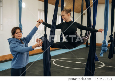 Controlling the twine exercise. Female instructor helping a child practice aerial yoga on hammock straps in a fitness studio Controlling the twine exercise. Female instructor helping a child practice aerial yoga on hammock straps in a fitness studio 139320494