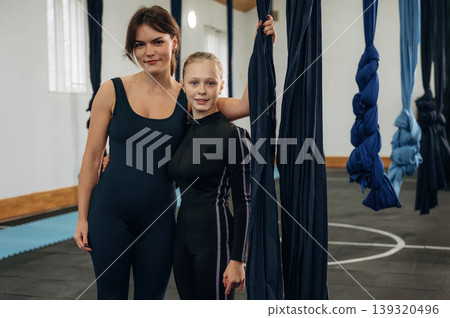 Embracing each other. Female instructor helping a child practice aerial yoga on hammock straps in a fitness studio Embracing each other. Female instructor helping a child practice aerial yoga on hammock straps in a fitness studio 139320496