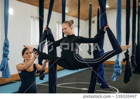 Controlling the twine exercise. Female instructor helping a child practice aerial yoga on hammock straps in a fitness studio Controlling the twine exercise. Female instructor helping a child practice aerial yoga on hammock straps in a fitness studio 139320497