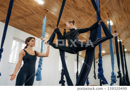 Controlling the twine exercise. Female instructor helping a child practice aerial yoga on hammock straps in a fitness studio 139320499