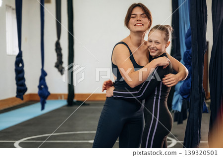 Embracing each other. Female instructor helping a child practice aerial yoga on hammock straps in a fitness studio Embracing each other. Female instructor helping a child practice aerial yoga on hammock straps in a fitness studio 139320501