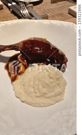 Roasted duck leg accompanied by creamy mashed potatoes and mushroom sauce on a white plate, set on a wooden table with cutlery in the background Roasted duck leg accompanied by creamy mashed potatoes and mushroom sauce on a white plate, set on a wooden table with cutlery in the background 139321666