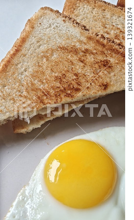 Close-up view of toasted bread slices with golden-brown crust alongside a sunny-side-up egg on a white plate, showcasing a simple breakfast arrangement Close-up view of toasted bread slices with golden-brown crust alongside a sunny-side-up egg on a white plate, showcasing a simple breakfast arrangement 139321674