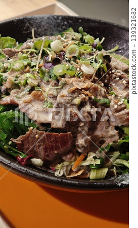 A vibrant salad featuring tender sliced beef, mixed greens, sesame seeds, and chopped green onions presented in a black bowl on an orange background A vibrant salad featuring tender sliced beef, mixed greens, sesame seeds, and chopped green onions presented in a black bowl on an orange background 139321682