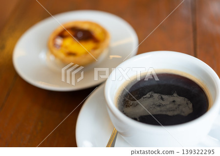 Hot black coffee in white cup with creamy foam served with custard tart dessert on wooden table warm cafe atmosphere soft focus and cozy morning break Hot black coffee in white cup with creamy foam served with custard tart dessert on wooden table warm cafe atmosphere soft focus and cozy morning break 139322295