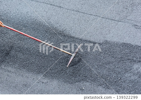 Worker using rake to spread asphalt evenly on road surface construction maintenance process texture detail infrastructure repair outdoor labor activity 139322299