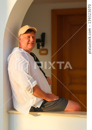 Adult man in a white shirt and cap sitting on a wide windowsill, enjoying the beautiful sunset, reflecting on life and tranquility in the golden hour 139323070