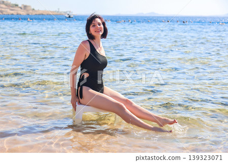 Elegant woman in a black swimsuit sitting on a plastic chair, enjoying the serene sea view, capturing the essence of summer relaxation and beach vibes Elegant woman in a black swimsuit sitting on a plastic chair, enjoying the serene sea view, capturing the essence of summer relaxation and beach vibes 139323071