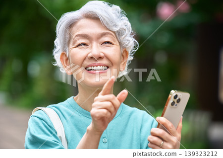 A close-up of a senior woman holding a smartphone and smiling with her finger raised. 139323212