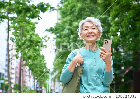 一位老婦人面帶微笑地抬頭看著窗外,手裡拿著智慧型手機,站在綠樹成蔭的街道上。 一位老婦人面帶微笑地抬頭看著窗外,手裡拿著智慧型手機,站在綠樹成蔭的街道上。 139324332
