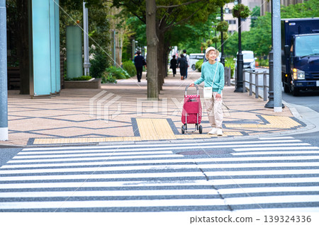 一位老婦人拉著購物車在十字路口散步。 一位老婦人拉著購物車在十字路口散步。 139324336