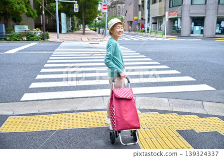 A senior woman with a shopping cart turns around and smiles at a crosswalk. A senior woman with a shopping cart turns around and smiles at a crosswalk. 139324337