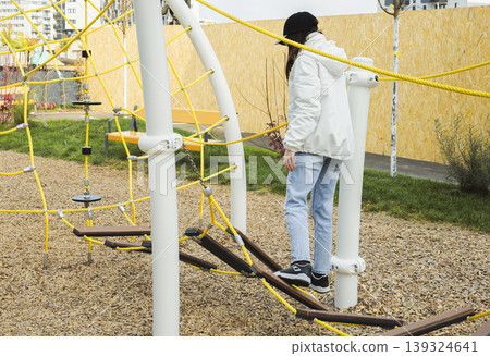 Young woman walks across a rope bridge in an outdoor playground, capturing the joy of simple challenges. Young woman walks across a rope bridge in an outdoor playground, capturing the joy of simple challenges. 139324641