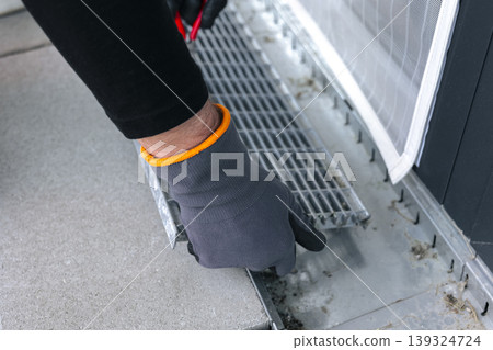 A worker wearing safety gloves removes a metal drain grate using pliers, illustrating a hands on drainage maintenance process 139324724