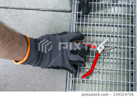 A person wearing safety gloves adjust a metal drain grate, highlighting practical outdoor repair and routine upkeep. 139324726