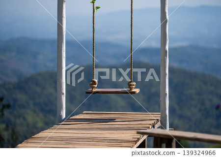 Vertical shot of beautiful white wooden swings decorated with floral ivy and wooden bridge located along mountain and cliff of flower field in North of Thailand. Vertical shot of beautiful white wooden swings decorated with floral ivy and wooden bridge located along mountain and cliff of flower field in North of Thailand. 139324966