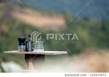 The coffee grinder, kettle, and coffee pot arranged on the table at the beautiful flower field view above the forest and mountains background The coffee grinder, kettle, and coffee pot arranged on the table at the beautiful flower field view above the forest and mountains background 139324971