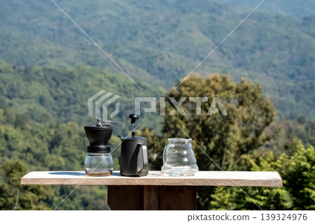 The coffee grinder, kettle, and coffee pot arranged on the table at the beautiful flower field view above the forest and mountains background The coffee grinder, kettle, and coffee pot arranged on the table at the beautiful flower field view above the forest and mountains background 139324976