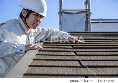一名男工人在檢查一棟獨立房屋的屋頂材料。 一名男工人在檢查一棟獨立房屋的屋頂材料。 139325068