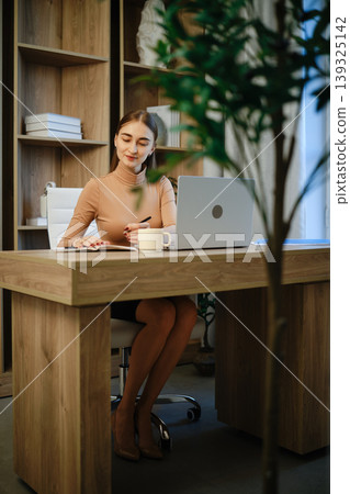Woman working at a wooden desk in a modern office space with a laptop and a cup of coffee Woman working at a wooden desk in a modern office space with a laptop and a cup of coffee 139325142