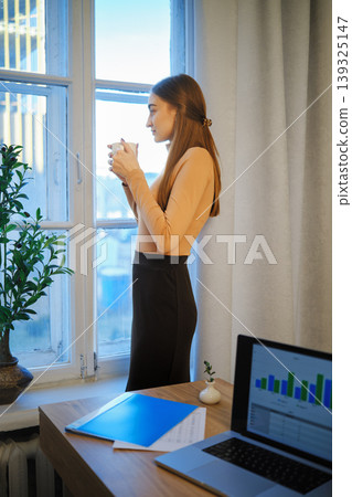 Woman standing by the window holding a cup while looking outside in an office setting Woman standing by the window holding a cup while looking outside in an office setting 139325147