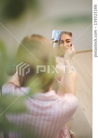 Woman is holding a mirror in front of her face while sitting indoors with plants around her Woman is holding a mirror in front of her face while sitting indoors with plants around her 139325166