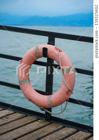 Rescue lifebuoy attached to pier railing above calm lake water, safety equipment at waterfront with scenic mountain view, travel concept 139325602