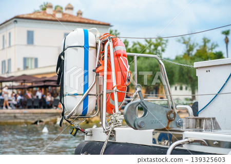 Lifebuoy and safety gear mounted on boat in Lake Garda marina, Italy, with waterfront cafe and people in background, nautical travel, safety concept Lifebuoy and safety gear mounted on boat in Lake Garda marina, Italy, with waterfront cafe and people in background, nautical travel, safety concept 139325603