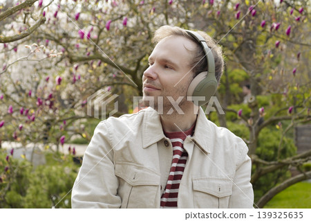 A smiling man with headphones relaxes in nature among blooming magnolias in a Japanese park, enjoying a digital detox. 139325635