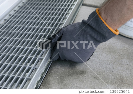 A worker wearing safety gloves removes a metal drain grate, illustrating a hands on drainage maintenance process A worker wearing safety gloves removes a metal drain grate, illustrating a hands on drainage maintenance process 139325641