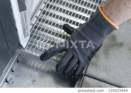 Close up of a hand in safety gloves adjusting a metal drain grate, highlighting practical outdoor repair and routine upkeep. 139325644