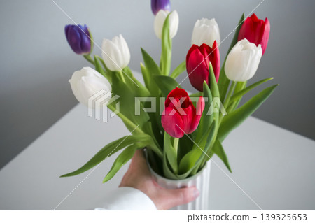 Hand holding a white ribbed vase with a colourful bouquet of tulips Hand holding a white ribbed vase with a colourful bouquet of tulips 139325653