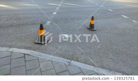 Two black traffic cones with orange reflective stripes stand on an asphalt road near a paved sidewalk. The markings on the street and the urban setting suggest temporary roadwork or a restricted area Two black traffic cones with orange reflective stripes stand on an asphalt road near a paved sidewalk. The markings on the street and the urban setting suggest temporary roadwork or a restricted area 139325781