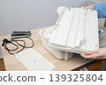 Person removing the outer casing of a white desktop printer scanner for maintenance and repair on a wooden desk, with detached parts, power cord and tools nearby in a tidy home office workspace. 139325804
