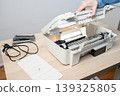 Person performing maintenance on an open white inkjet printer scanner on desk, showing internal components, power cable and removed parts, illustrating repair, troubleshooting and office technology. 139325805