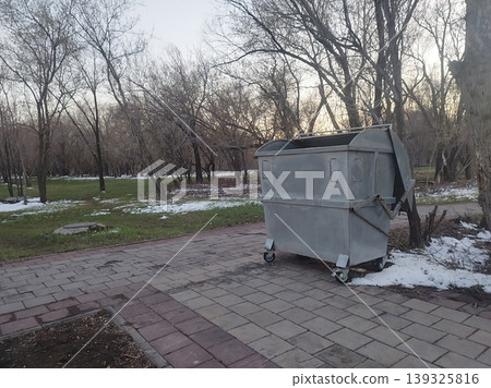 Large metal garbage container stands on a paved park walkway. In the background, bare trees and patches of melting snow on green grass signal the transition from winter to spring in an urban park Large metal garbage container stands on a paved park walkway. In the background, bare trees and patches of melting snow on green grass signal the transition from winter to spring in an urban park 139325816