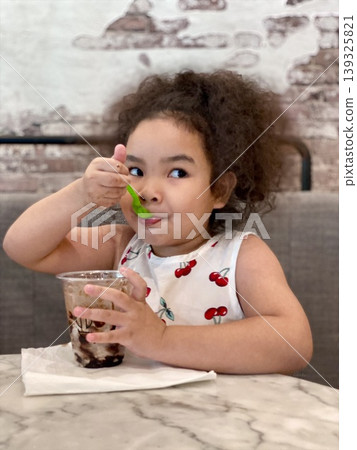Portrait of cute Asian little girl with curly hair eating chocolate ice cream in a glass with a green spoon in her hand at a cafe Portrait of cute Asian little girl with curly hair eating chocolate ice cream in a glass with a green spoon in her hand at a cafe 139325821
