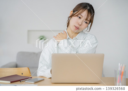 A woman suffering from stiff shoulders while working on her computer in the living room. A woman suffering from stiff shoulders while working on her computer in the living room. 139326568