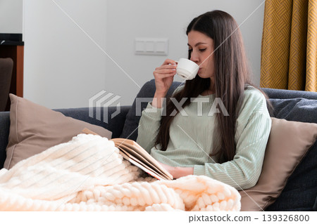 Peaceful morning lifestyle scene with young woman reading a book at home, wrapped in cozy blanket and drinking coffee. Relaxation, self care, hygge concept, calm mood, comfortable home interior and 139326800
