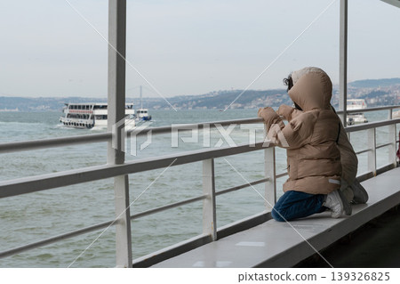 Children sit on waterfront railing in Istanbul looking at ferry crossing Bosphorus, distant coastline. Travel lifestyle, family tourism, urban exploration, childhood experience, transport journey Children sit on waterfront railing in Istanbul looking at ferry crossing Bosphorus, distant coastline. Travel lifestyle, family tourism, urban exploration, childhood experience, transport journey 139326825