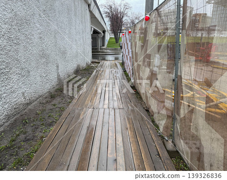 Wooden walkway along construction fence, concrete wall with wet surface in urban outdoor environment. Infrastructure maintenance, urban development, pedestrian path, city environment, construction Wooden walkway along construction fence, concrete wall with wet surface in urban outdoor environment. Infrastructure maintenance, urban development, pedestrian path, city environment, construction 139326836