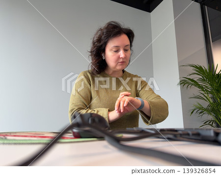 Woman checking smartwatch at desk in modern office interior with computer equipment, plant. Business workflow, time management, corporate environment, productivity concept, workplace routine Woman checking smartwatch at desk in modern office interior with computer equipment, plant. Business workflow, time management, corporate environment, productivity concept, workplace routine 139326854