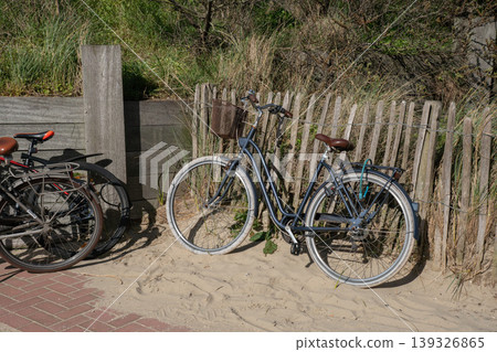 Bicycles parked on sandy beach near wooden fence, dune grass under natural daylight. Urban mobility, eco transport, outdoor lifestyle, coastal travel, sustainable transportation content Bicycles parked on sandy beach near wooden fence, dune grass under natural daylight. Urban mobility, eco transport, outdoor lifestyle, coastal travel, sustainable transportation content 139326865