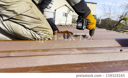 Male hands in gloves use cordless screwdriver to fasten roofing sheets in house roof. Young worker secures screws into surface performing careful installation work during residential construction Male hands in gloves use cordless screwdriver to fasten roofing sheets in house roof. Young worker secures screws into surface performing careful installation work during residential construction 139328165