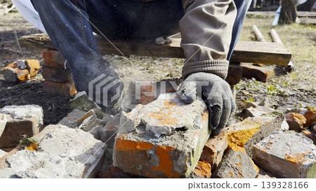 Male hands in protective gloves uses hammer to remove old cement from used bricks during renovation work. The process involves carefully cleaning brick surface to prepare it for reuse in construction. Male hands in protective gloves uses hammer to remove old cement from used bricks during renovation work. The process involves carefully cleaning brick surface to prepare it for reuse in construction. 139328166
