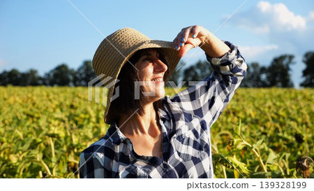 Happy smiling female farmer looks into sunflower field and enjoys harvest. Portrait of adult beautiful agronomist in straw hat with yellow plantation at background. Concept of agricultural business Happy smiling female farmer looks into sunflower field and enjoys harvest. Portrait of adult beautiful agronomist in straw hat with yellow plantation at background. Concept of agricultural business 139328199