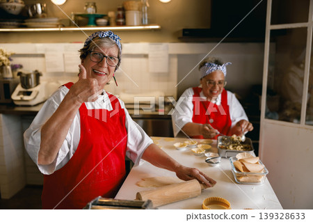 Senior woman in red apron giving thumbs up while rolling dough in bright kitchen Senior woman in red apron giving thumbs up while rolling dough in bright kitchen 139328633
