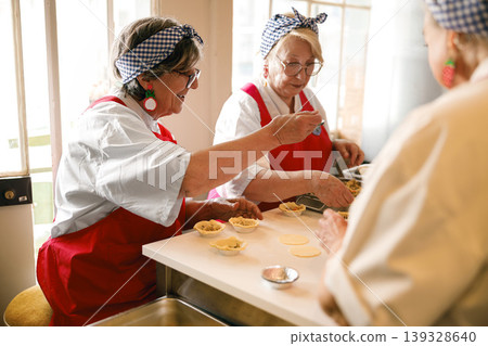 Three senior women filling pastry tarts together in a bright culinary workshop Three senior women filling pastry tarts together in a bright culinary workshop 139328640
