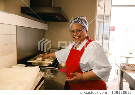 Happy senior woman in red apron standing in kitchen while preparing food Happy senior woman in red apron standing in kitchen while preparing food 139328652
