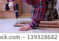 A worker in a plaid shirt operates a screwdriver on a disassembled wooden bed on a light floor in a home room, fixing fasteners on the side board of a light wood bed 139328682